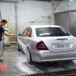 A young man using a hose to wash a silver car inside an indoor car wash facility.